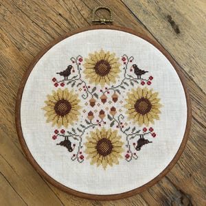 Puede incluir: Un bastidor de bordado circular de madera que contiene un patrón de punto de cruz sobre tela blanca. El diseño presenta cuatro girasoles amarillos con centros marrones, dispuestos simétricamente, intercalados con pequeñas bellotas marrones, bayas rojas y cuatro pájaros negros.