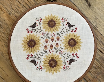 May include: A circular wooden embroidery hoop containing a cross-stitch pattern on white fabric. The design features four yellow sunflowers with brown centers, arranged symmetrically, interspersed with small brown acorns, red berries, and four black birds.