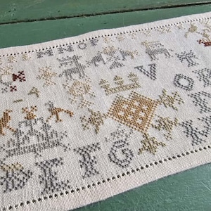 Könnte beinhalten: Ein handgesticktes Stickmuster mit cremefarbenem Hintergrund. Das Stickmuster zeigt verschiedene gestickte Motive, darunter Vögel, Hirsche und geometrische Formen, zusammen mit dem Alphabet und Zahlen. Die Farbpalette umfasst Braun-, Grau- und Goldtöne.