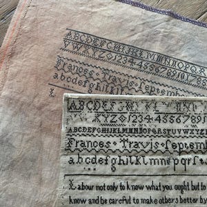 Könnte beinhalten: Nahaufnahme antiker Nadelstickerei-Sampler mit schwarzen Buchstaben und Zahlen auf beigem Stoff. Der Text enthält den Namen "Frances Travis" und das Datum "September".