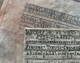 May include: Close-up of antique needlework samplers with black stitched lettering and numbers on a beige fabric. The text includes the name "Frances Travis" and the date "September".