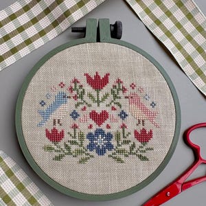 May include: A circular embroidery hoop containing a linen fabric with a cross-stitch pattern featuring a central red heart, a blue flower, two birds in blue and pink, and various red and green floral motifs. The hoop is dark green plastic.