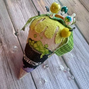 May include: A cone-shaped fabric pincushion featuring a cross-stitched black cauldron with green bubbling liquid and the word POTION. The top is adorned with green fabric, felt, and several decorative glass beads and small black bat charms.