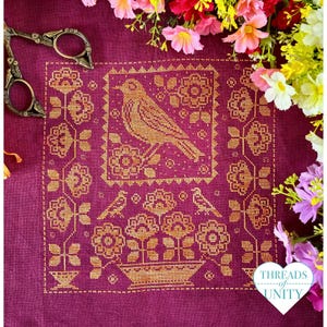 Peut inclure: Un tissu en lin magenta profond orné d'une broderie au point de croix dorée représentant un oiseau perché au-dessus d'un panier de fleurs, entouré d'une bordure florale. Une paire de ciseaux en métal de style ancien repose sur le tissu.