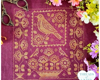 May include: A deep magenta linen fabric featuring a gold cross-stitch embroidery of a bird perched above a floral basket, surrounded by a floral border. A pair of vintage-style metal scissors rests on the upper left corner of the fabric.