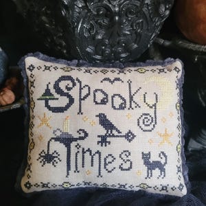 Könnte beinhalten: Dekoratives quadratisches Kissen mit den Worten "Spooky Times" in dunkelblauer Stickerei. Das Design umfasst einen Hexenhut, eine Krähe, eine Spinne, eine Katze, Sterne und einen Mond. Das Kissen hat einen hellen Hintergrund und einen dunkelblauen Fransenrand.