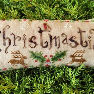 May include: A rectangular beige fabric cushion featuring cross-stitch embroidery. The word Christmastide is stitched in dark brown thread, accompanied by two brown reindeer, green holly leaves, red berries, and white snowflake motifs.