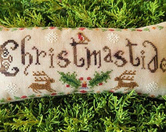 May include: A rectangular beige fabric pillow featuring cross-stitch embroidery. The word Christmastide is stitched in dark brown thread, accompanied by two brown reindeer, green holly leaves, red berries, and white snowflake motifs.