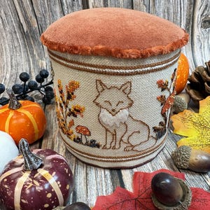 May include: A cylindrical pincushion featuring a rust-coloured suede top with a fuzzy trim. The sides are made of beige woven fabric embroidered with a fox, small mushrooms, and autumn foliage in shades of orange, brown, and yellow.