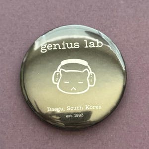 May include: A round, glossy button with the text "genius lab" in white. Below the text is a white illustration of a cat wearing headphones. The button also includes the text "Daegu, South Korea est. 1993".