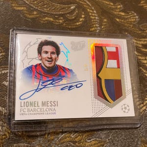 May include: A Lionel Messi FC Barcelona UEFA Champions League trading card in a protective plastic case. The card features a portrait of Messi, a blue ink autograph, and a 1/1 serial number. A fabric patch with the team colours is embedded on the right.