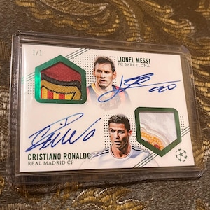 May include: A 1/1 sports trading card featuring Lionel Messi of FC Barcelona and Cristiano Ronaldo of Real Madrid CF. The card includes two player portraits, two blue ink autographs, and two embedded fabric jersey patches in green frames.