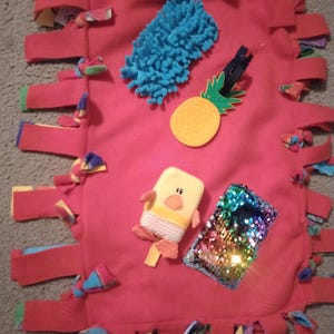 May include: A red fleece sensory blanket with various textures and shapes. Includes a blue textured item, a yellow pineapple, a small plush duck, and a reversible sequin patch. The blanket's edges are finished with colorful knotted strips.
