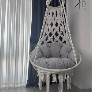 Puede incluir: Una silla colgante de macramé blanca con un cojín gris. La silla está suspendida de una cuerda y presenta un diseño de patrón de diamantes. La silla tiene flecos en la parte inferior y está diseñada para uso en interiores.