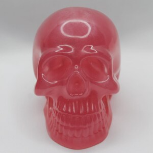 May include: A shiny, red skull ornament. The skull features detailed eye sockets, a nasal cavity, and a full set of teeth. The smooth surface reflects light, giving it a polished look. The skull is a decorative item.