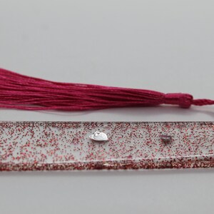 May include: A clear bookmark with red glitter and small embellishments, including a heart-shaped charm. A matching magenta tassel is attached to the top. The bookmark is a decorative accessory for books and journals.