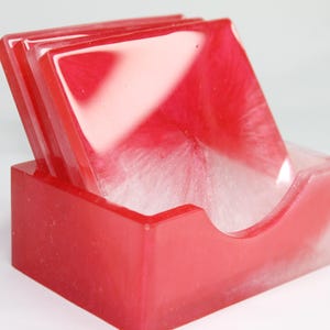 Puede incluir: Un juego de cuatro posavasos cuadrados en un diseño de resina roja y blanca, exhibidos en un soporte a juego. Los posavasos tienen un efecto veteado, con una mezcla de tonos rojos y blancos. El soporte tiene un recorte curvo.