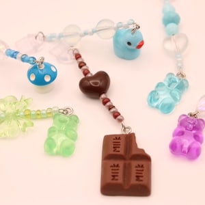 May include: A collection of colorful beaded charms with various pendants. Charms include a blue mushroom, a green gummy bear, a chocolate bar, a heart, and a purple gummy bear. The charms are connected by silver-colored metal rings.