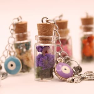May include: Several small glass bottle keyrings filled with dried flowers in different colours. Each bottle has a cork stopper and a silver chain. Some keyrings feature an evil eye charm or a dragonfly charm.