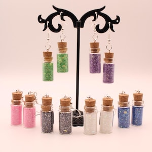 May include: Earrings featuring small glass bottles filled with colorful glitter. The earrings have cork stoppers and silver-tone hooks. Colors include pink, green, purple, silver, white, and blue. The earrings are displayed on a black metal stand.