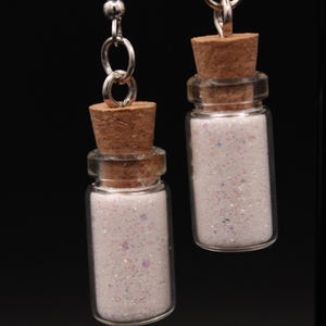 May include: A pair of dangle earrings featuring small glass bottles filled with white glitter. Each bottle is topped with a cork and suspended from a silver-tone hook. The glitter has iridescent specks.