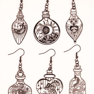 May include: Six black metal earrings with intricate designs. The earrings feature various shapes, including bottles and a heart, adorned with celestial and mystical motifs such as moons, suns, eyes, and mushrooms. Each earring has a hook for easy wear.