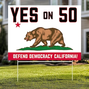 May include: A white yard sign with the text "YES ON 50" in black, a red star, and a brown bear illustration. The sign also includes the text "DEFEND DEMOCRACY CALIFORNIA!" in red. The sign is displayed on a green lawn.