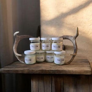 Puede incluir: Varios frascos de vidrio con tapas blancas, etiquetados como "TALLOW", dispuestos sobre una superficie de madera rústica, junto a una cornamenta de ciervo. Los frascos contienen una sustancia cremosa, creando una estética natural.