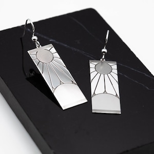 Puede incluir: Pendientes plateados con diseño rectangular y patrón de sol. Los pendientes tienen un acabado pulido y se muestran sobre una superficie negra. Cierre de gancho.