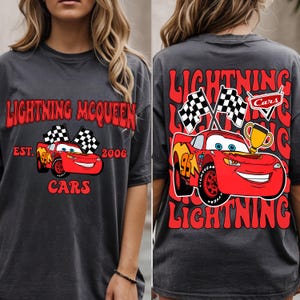 Può includere: T-shirt grigio scuro con Lightning McQueen del film Cars. La parte anteriore mostra "Lightning McQueen" e "Cars", con un'auto da corsa rossa e bandiere a scacchi. Il retro ripete "Lightning" e presenta l'auto, le bandiere e un trofeo.