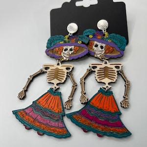 Op de afbeelding: Houten Day of the Dead oorbellen met een skeletontwerp. De oorbellen hebben een skeletkop met een hoed met bloemaccenten en een kleurrijke gelaagde rok. De oorbellen zijn bevestigd aan een zwarte displaykaart.