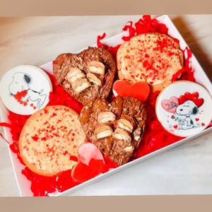 May include: A white box filled with red paper and assorted biscuits. The biscuits include heart-shaped brownies topped with sweets, round biscuits with red sprinkles, and biscuits decorated with Snoopy and hearts. A Valentine's Day gift.