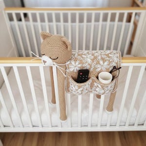 Op de afbeelding: Een gehaakte katvormige organizer in bruin met witte accenten, ontworpen om aan een wieg te hangen. De organizer heeft vakken voor items zoals een afstandsbediening, een bril en een kleine beker. De kat heeft gesloten ogen en witte snorharen.