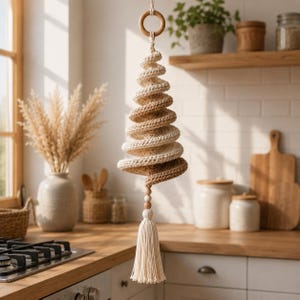 May include: A hanging macrame decoration featuring a spiral, tree-like shape crafted from alternating cream and tan cotton cord. The piece is suspended from a wooden ring at the top and finished with two small wooden beads and a long cream tassel.