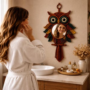 May include: A decorative owl-shaped mirror with a round mirror in the center. The owl is made of woven fibers in shades of brown, red, green, and yellow. The mirror hangs on a beige wall above a bathroom vanity.