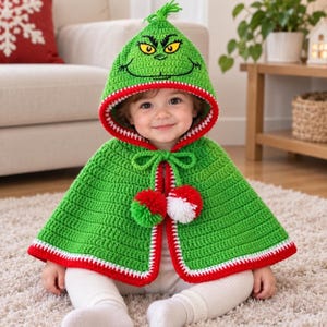 Op de afbeelding: Een groene gehaakte cape met capuchon, voorzien van een karaktergezicht met gele ogen en een brede grijns op de kap. De cape heeft een rood-witte rand, een striksluiting bij de hals en twee grote pompons in rood, wit en groen aan de voorzijde.