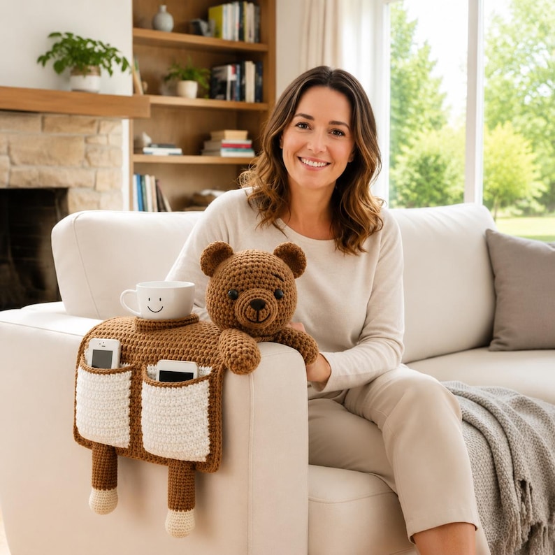 Puede incluir: Un organizador de brazo de sof&aacute; de ganchillo marr&oacute;n y crema con forma de oso. Cuenta con dos bolsillos para tel&eacute;fonos, un portavasos con una cara sonriente y un peluche de oso a juego. Los brazos del oso est&aacute;n sobre el brazo del sof&aacute;.