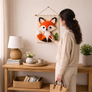 May include: A handmade, orange, white, and black fox wall hanging. The fox is crocheted and mounted on a cream-colored fabric with a wooden dowel for hanging. The fox has a friendly expression and is surrounded by green leaves.
