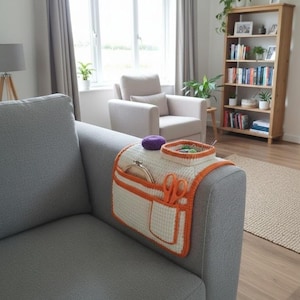 Peut inclure: Un organiseur de bras de canapé en crochet blanc et orange avec des poches, contenant des ciseaux, un cercle à broder et un petit panier. Un coussin à épingles violet est posé dessus. L'organisateur est placé sur un bras de canapé gris.