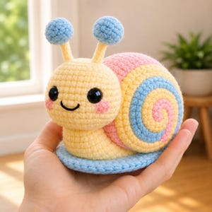 Puede incluir: Un juguete de caracol tejido a ganchillo en amarillo, rosa y azul. El caracol tiene una cara sonriente con mejillas rosadas, ojos negros grandes y antenas azules. La concha es una espiral de amarillo, rosa y azul. El caracol está sobre una base azul.