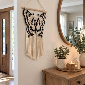 May include: A macrame wall hanging shaped like a butterfly, crafted from cream and black cord. The butterfly features intricate knotted patterns, two antennae, and long, flowing fringe tassels hanging from the bottom, suspended by a wooden dowel.