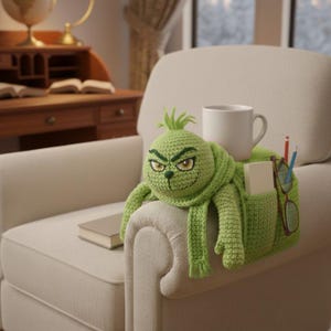 May include: A green crocheted Grinch-themed armrest organizer with a smiling face, scarf, and pockets. A white mug, pencils, glasses, and a notepad are in the pockets. A book rests on the armrest.