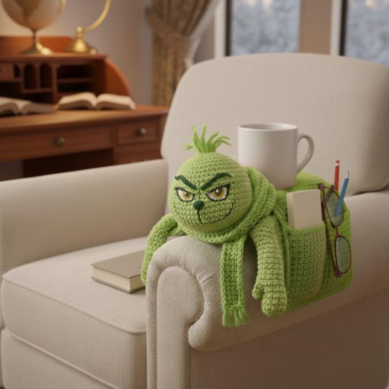 May include: A green crocheted Grinch-themed armrest organizer with a smiling face, scarf, and pockets. A white mug, pencils, glasses, and a notepad are in the pockets. A book rests on the armrest.
