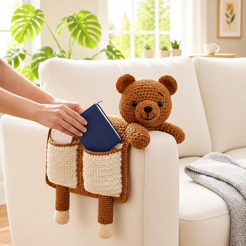Puede incluir: Un oso de peluche marr&oacute;n tejido a ganchillo con un organizador de bolsillo adjunto a un brazo de sof&aacute; blanco. El organizador tiene dos bolsillos con la parte superior de color crema y la inferior marr&oacute;n. Se est&aacute; colocando un libro azul en uno de los bolsillos.