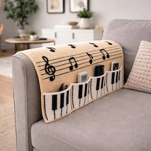 Könnte beinhalten: Beige gehäkelte Sofalehnenabdeckung mit schwarzen Noten und einem Klaviertasten-Design. Verfügt über drei Taschen für Fernbedienungen und andere kleine Gegenstände. Ein dekoratives und funktionales Wohnaccessoire.