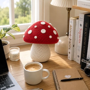 May include: A handcrafted crochet mushroom with a vibrant red cap featuring white circular spots and a cream-colored stem. The mushroom is constructed with a dense, textured yarn stitch pattern, creating a rounded top and a cylindrical base.