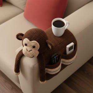 May include: A brown crochet monkey armrest with a light tan face and ears. The armrest has two pockets with blue and tan stripes and holds two remote controls. A white mug of coffee sits on top.