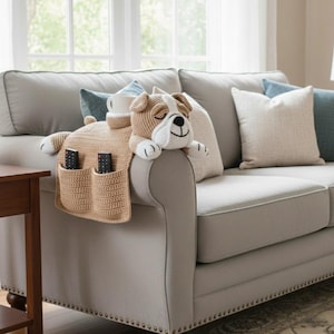 Puede incluir: Organizador de reposabrazos de sofá de ganchillo beige con forma de bulldog. El organizador tiene dos bolsillos para mandos a distancia y un espacio para una taza. El bulldog tiene la cara blanca, orejas marrones y está sobre el brazo de un sofá gris.