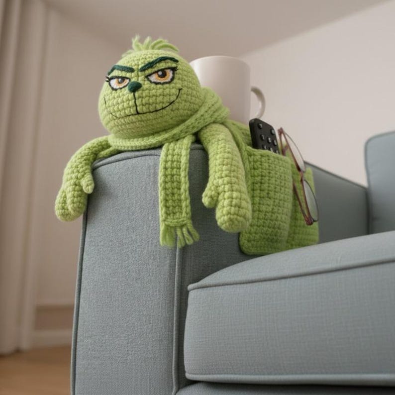 May include: A green crocheted Grinch-themed couch caddy with a scarf, arms, and a pocket for a remote and glasses. The caddy is placed on the arm of a light blue couch, with a white mug visible in the background.