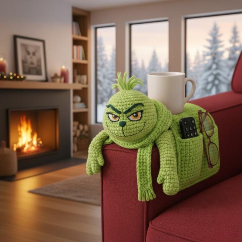 May include: A green crocheted Grinch-themed couch armrest organizer with a smiling face, arms, and scarf. It features pockets for a remote and glasses, and holds a white mug. The armrest is on a red couch.